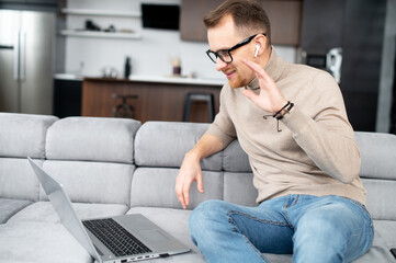 Front view of cheerful fit young bearded hipster man in glasses and wireless earphones sitting on the couch, waving at laptop, video-calling with coworkers online, male freelancer talking on webcam