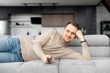 Young bearded hipster man lying down on the couch in the living room, pointing a tv remote on the camera, switching channels, happy with a television program, watching movie, series, chilling at home