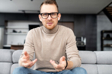 Portrait of intelligent young bearded hipster man looking at camera, wearing glasses and wireless earphones, sitting on a couch in the living room and gesturing. The view while video-calling concept