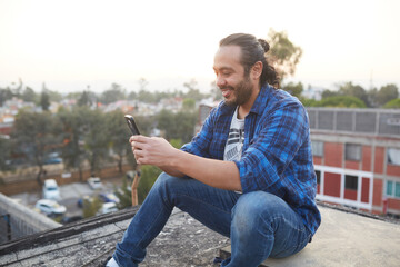 30 year old man sitting on the roof of a building watching the sunset and 30-year-old man sitting on his back on the rooftop of a building watching the sunset and browsing on his smart cell phone