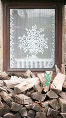 Visillo de encaje blanco con motivo floral en ventana junto a pila de troncos de leña en fachada...