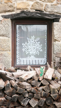 Visillo de encaje blanco con motivo floral en ventana junto a pila de troncos de le&ntilde;a en fachada de piedra