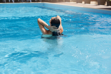 Young woman posing backwards while standing in blue water and touching her wet dark hair, female swims in swimming pool, back view of girl relaxing in drainage basin.