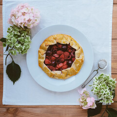 Delicious freshly baked vegan strawberry and cherry galette on wooden rustic background with hydrangea flowers, top view. Sweet food, summer dessert.