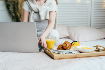 Obraz na płótnie Canvas An unrecognizable millennial woman is sitting on the bed and typing on a laptop, next to a tray with breakfast. Home office vibes.