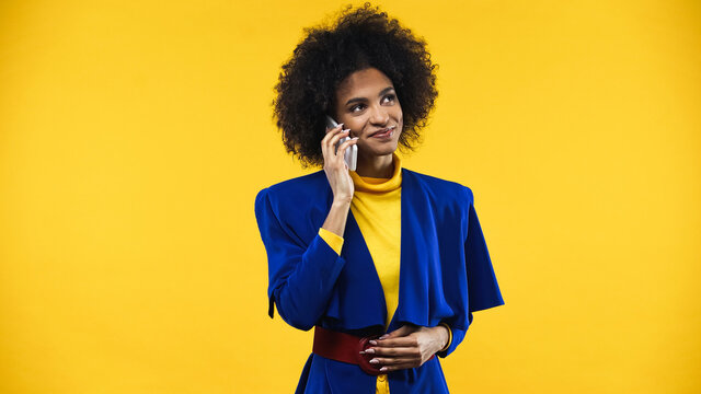 Smiling African American Woman In Blue Jacket Talking On Smartphone Isolated On Yellow