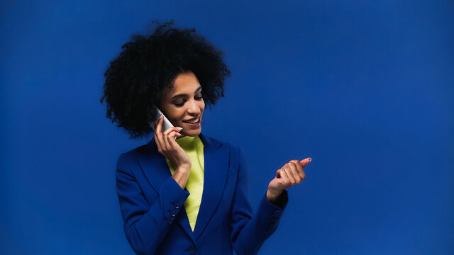 Smiling african american woman talking on smartphone and looking at nails isolated on blue