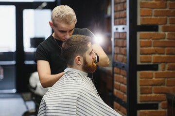 Making haircut look perfect. Young bearded man getting haircut by hairdresser while sitting in chair at barbershop