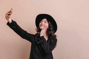 Romantic asian woman in coat taking selfie. Studio shot of inspired chinese girl in hat.