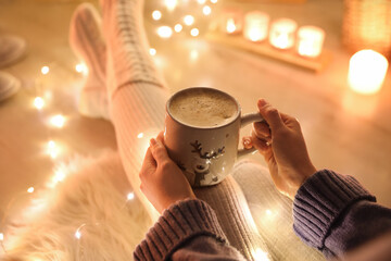 Woman with cup of drink on floor against blurred Christmas lights, closeup