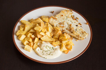 Dish with a hake fillet with garlic and parsley accompanied by aioli sauce and potatoes on a dark wooden table. Food and plating.