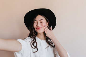 Blissful asian girl taking selfie with closed eyes. Studio shot of blithesome japanese model in black hat.