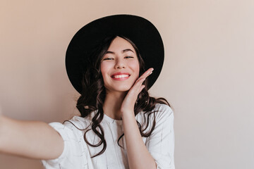 Jocund asian girl in hat laughing at camera. Glamorous chinese woman taking selfie.