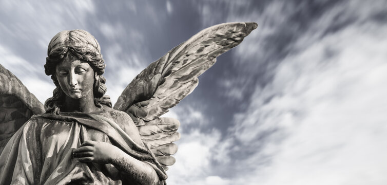 Sad guardian angel sculpture with open wings isolated with blurred white clouds dramatic sky. Sorrow angel sad expression sculpture with eyes down and hand in front of chest. Desaturated picture.