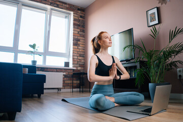 Fototapeta na wymiar Low-angle view of happy redhead young woman wearing sportswear is meditating in lotus position sitting on yoga mat near laptop. Peaceful red-haired lady holding hands in Namaste pose near chest.
