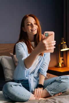 Charming redhead young woman taking mobile selfie photo on cellphone at cozy bedroom. Happy lady wearing home clothes making selfie on smartphone. Closeup portrait of cheerful female in bed.