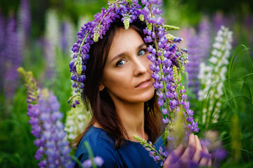 Large portrait of a woman in a wreath on a lupine field. A meadow of purple flowers in summer. A girl with long hair holds a bouquet of lupines against the background of nature.