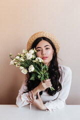 Stylish asian girl in straw hat holding white eustomas. Front view of lovely curly woman with flower bouquet on beige background.