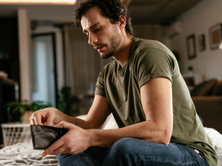 Young man checking empty wallet, no money. Unemployed sad man sitting at home thinking about money.