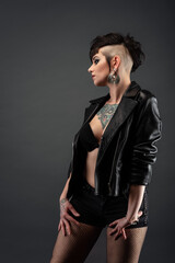 Fototapeta premium Attractive tattooed young woman, in profile, with a punk hairstyle, wearing a bra, leather jacket and fishnet stockings