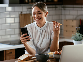 Beautiful woman eating sandwich. Smiling woman using the phone in the kitchen.