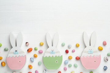 Candies and bunny figures in protective masks on white wooden table, flat lay with space for text. Easter holiday during COVID-19 quarantine