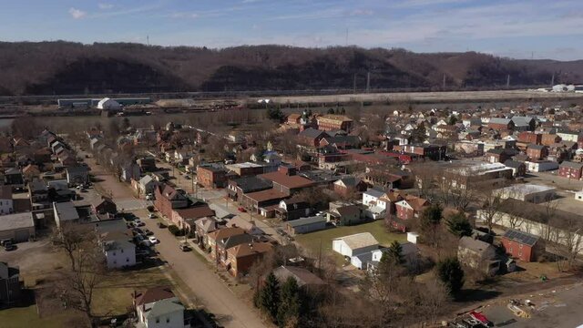A cinematic slow orbiting aerial establishing shot of a typical Pennsylvania small town in late winter. Pittsburgh suburbs. Factory on the Ohio River in the distance.  	