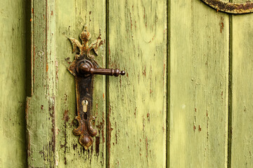 Old rusty door handle on green door. Detail of rustic old vintage and rusty grungy iron door handle installed on an ornamented old heavy and massive medieval green wooden door with peeling paint.