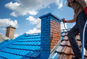Professional using special equipment to paint chimney in blue color. Half roof is already painted....