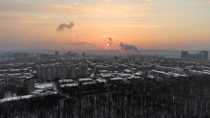 Aerial view Kharkiv city Pavlove Pole district. Smoke from pipes above winter dark forest, multistorey houses and snowy orange sunset sky