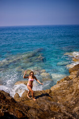 Young beautiful woman in a swimsuit stands on a rocky beach of the Mediterranean Sea. The concept of sea recreation. Selective focus