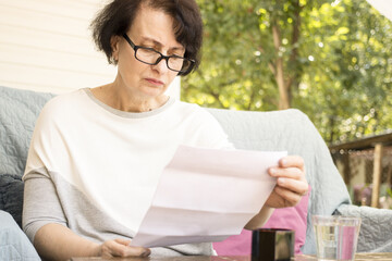 Serious worried senior woman in eyeglasses reading paper document or postal letter feeling frustrated by unexpected news sitting on sofa on home terrace. Received bank account balance or bad notice.