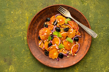 A healthy salad of sliced bloody oranges, purple onions, olives and pine nuts with olive oil dressing on a brown plate against an olive concrete background. Vegan recipes.