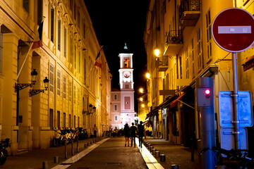 Three young people walk through old town Vieux Nice, France, at night with the Rusca Palace tower seen slightly blurred in the distance.