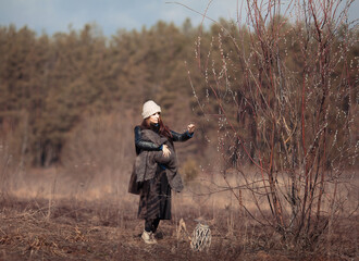 Fototapeta premium a girl in a knitted hat and fur coat, a long skirt touches a willow branch with one hand, and with the other hand holds a small child wrapped in a knitted scarf 