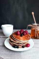 Pancakes with berries and honey on a white plate, wooden spoon, jar, coffee cup