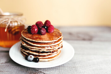 Juicy pancakes with berries and honey on a white plate, spoon, jar, wooden table