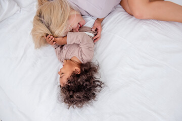 Young Caucasian blond mother tickles her little African American daughter. Lie on white bed, having...