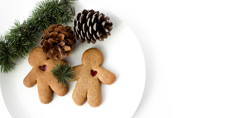 A pair of gingerbread lovers with hearts on white plate with a branch of a christmas tree and a pine cone 