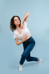 Cute contemporary dancer poses in front of blue studio background. She's crossing her legs in a lunge forward. Bending arms in elbows and slightly tilting her body.