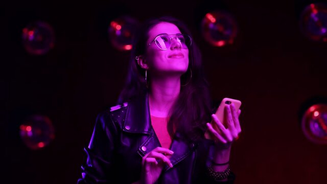 Close up. Slow motion. Beautiful brunette girl in club atmosphere. Happy female in sunglasses standing in room in the pink and blue neon light, texting on cellphone and smiling.