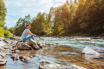 Young man relaxing by mountain river enjoying natural landscape. Traveler backpacker sitting on rock. Summer trip