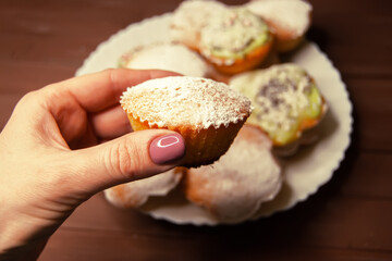 A cupcake baked at home in the girl's hand, sprinkled with powdered sugar, against the background of other homemade cakes, close-up. Leisure at home. Step-by-step recipe.