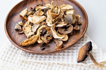Composition in natural light. Dried wild mushrooms of various shapes in a dark wood plate, and a large olin mushroom nearby. On a white kitchen towel