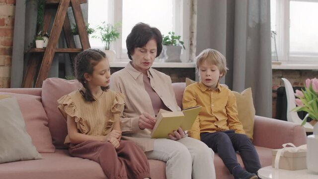 Medium PAN with slowmo of kind grandmother reading book to her two grandchildren listening, sitting together on comfy soft couch in living room