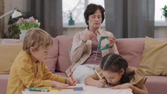 Slow-motion panning medium shot of playful kids drawing at table while their grandmother sitting on sofa in background knitting