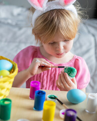 Easter holiday craft at home. Little girl in Easter bunny ears painting colored eggs.