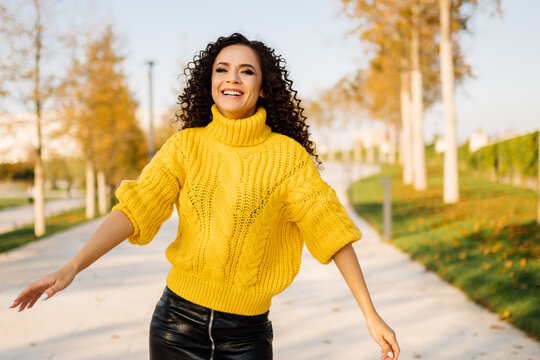 On the light alley of the park with yellow trees at the edges stands in a yellow sweater and a black skirt laughing curly brunette. High quality photo