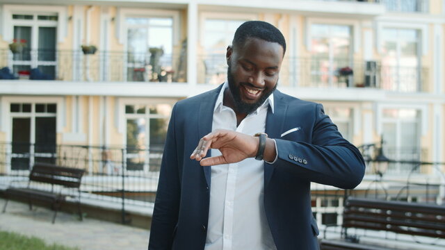 Smiling afro guy checking time outdoors. Businessman standing on urban street.