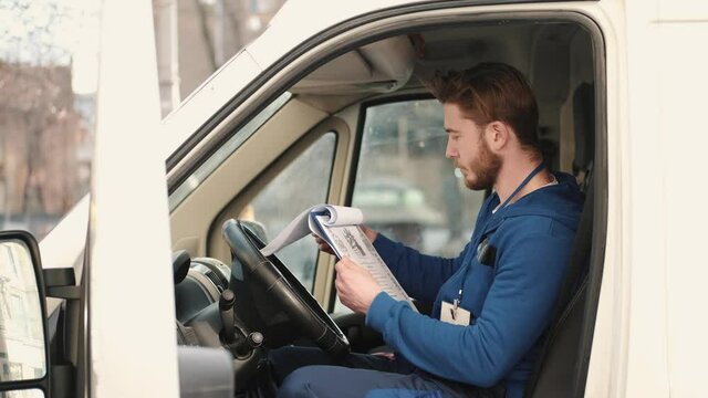 Delivery service man sitting in van and writing documents on clipboard before delivering a package to customer. Checking list, working process.